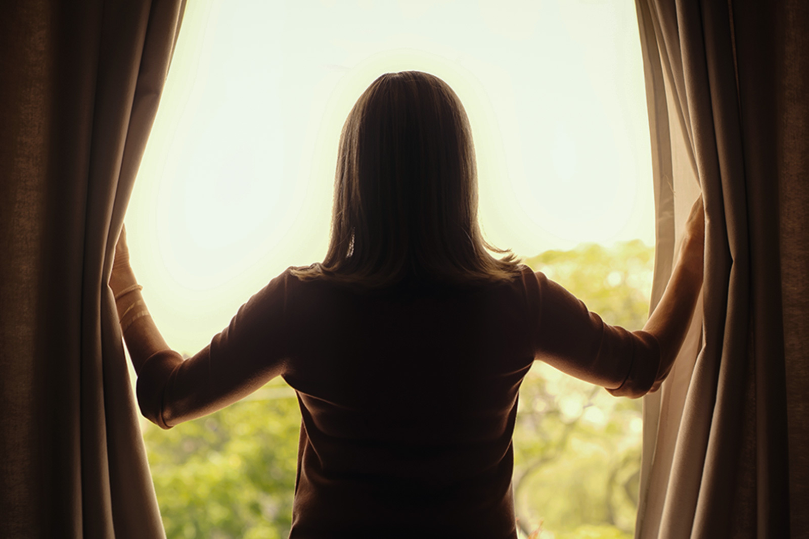 a woman in silhouette draws back the curtains and looks...