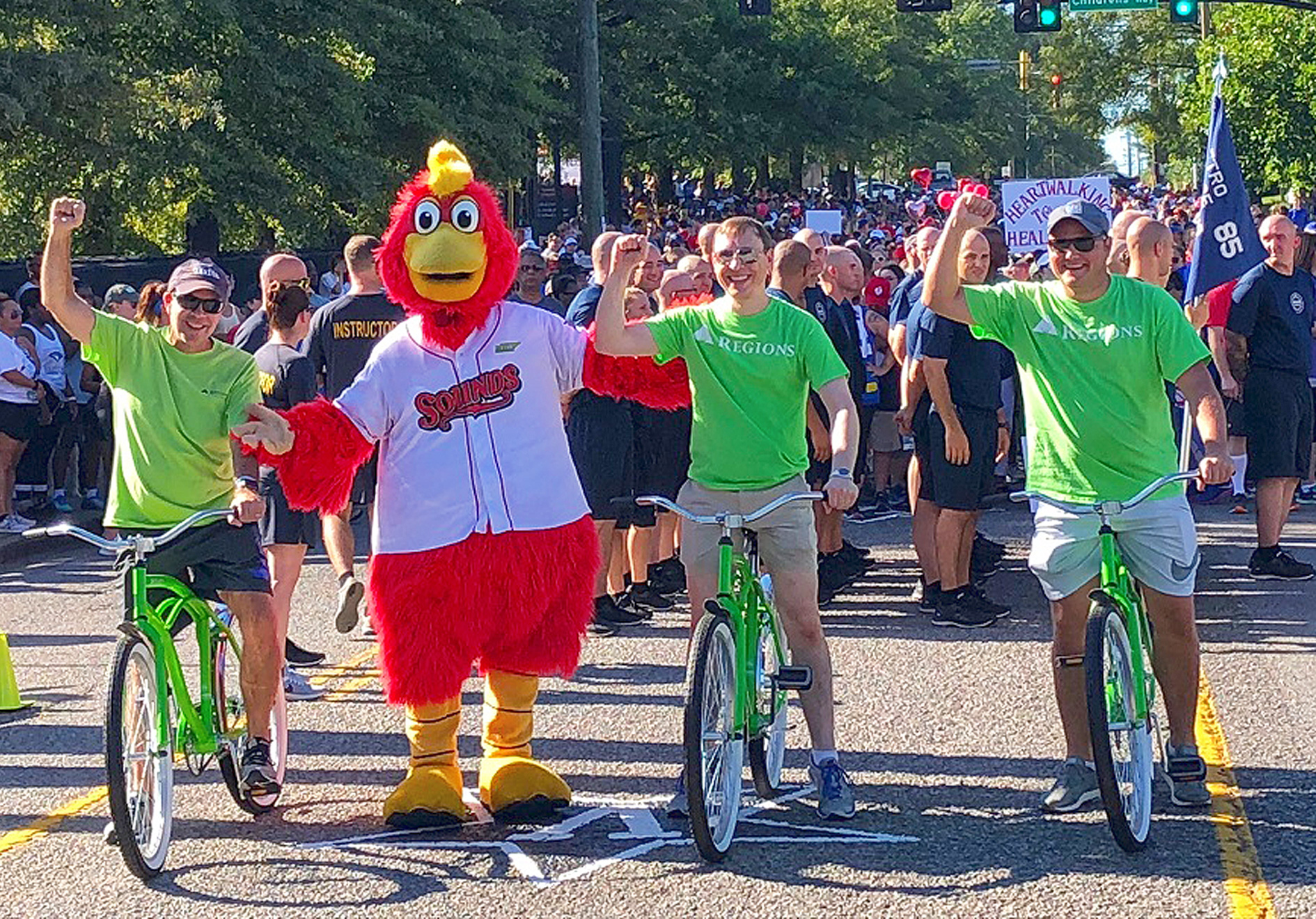 three men on bikes, with person standing in chicken mascot costume
