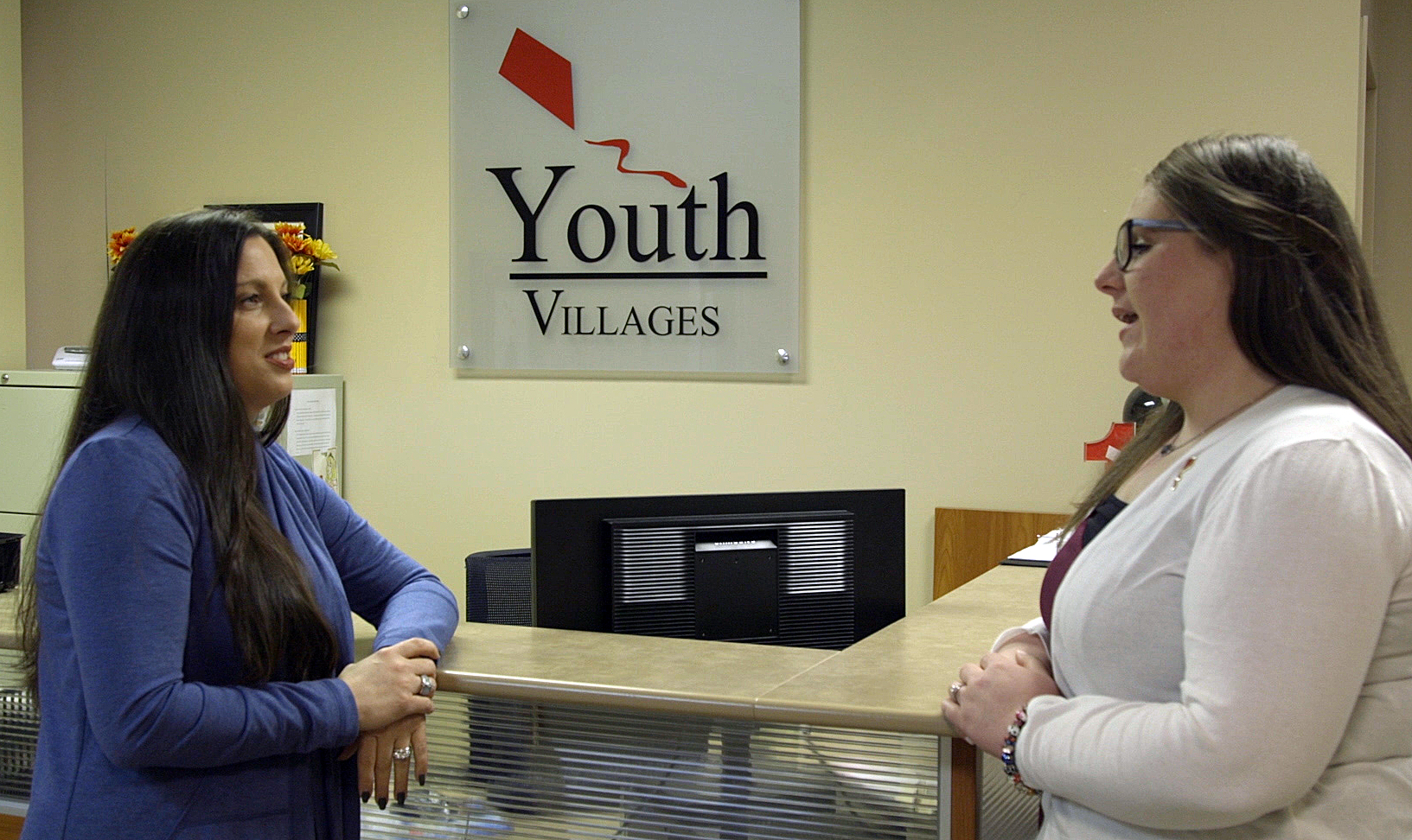 two women standing and talking, with sign in background that says Youth Village